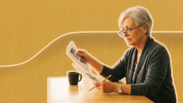 A silver-haired woman in her seventies reads a printed page at a kitchen table, a pencil in one hand and a mug of coffee beside her.